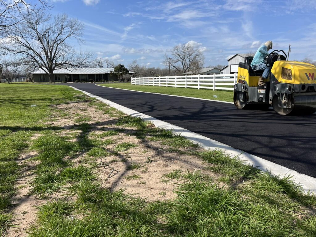 Paving crew works on a dark asphalt driveway beside a sidewalk